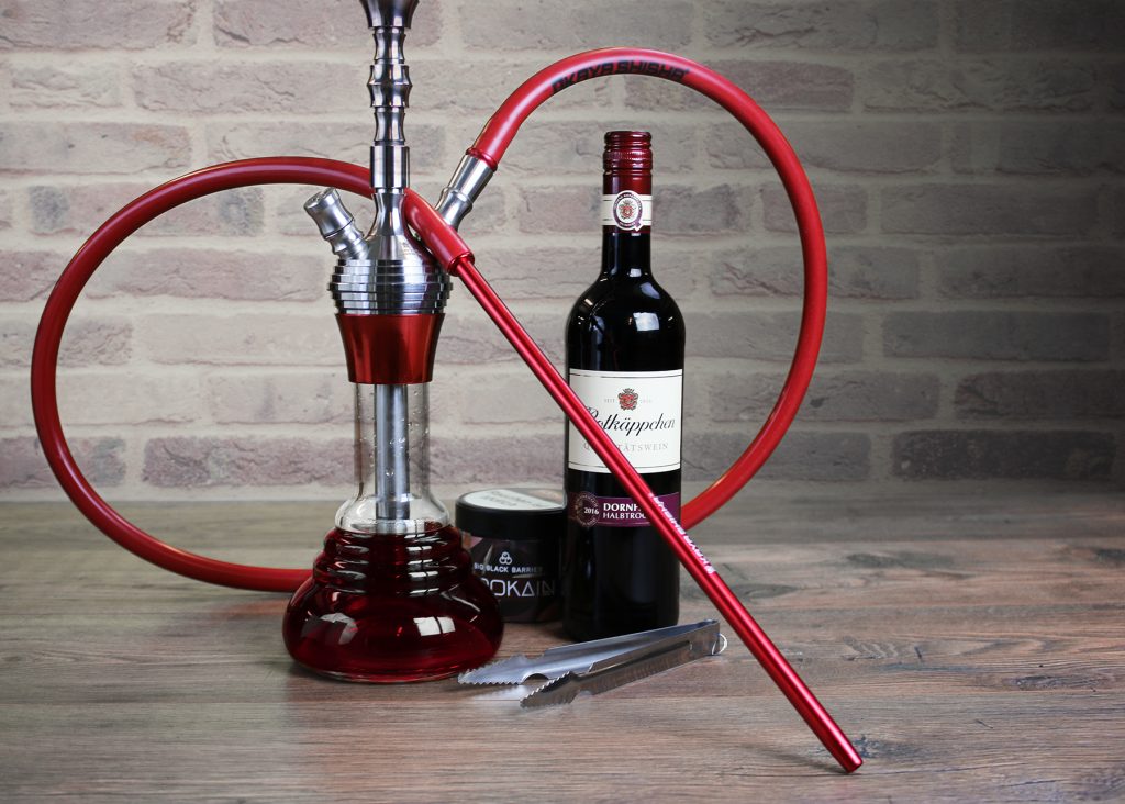 Eine mit Wein gefüllte Shisha, eine Rotweinflasche, eine Tabakdose und eine Kohlezange auf einem Holztisch vor einer Backsteinwand.