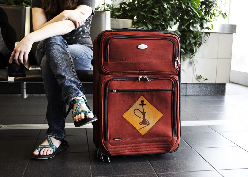 Sitzende Frau im Anschnitt, die am Flughafen mit dem Pass in der Hand wartet. Vor ihr ein roter Koffer mit gelbem Shisha Warnzeichen.
