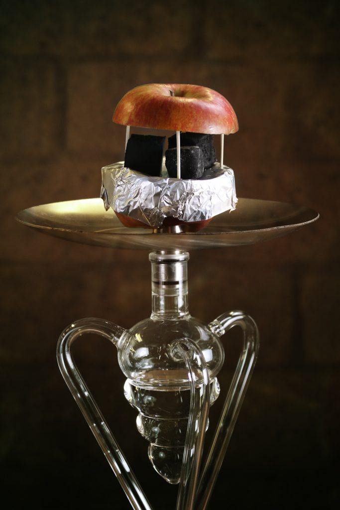 Nahaufnahme eines Apfel Fruchtkopfes auf einer Shisha. Die untere Hälfte ist mit Alufolie bedeckt. Darauf liegen 3 Stück unangezündete Kohle. Die obere Apfelhälfte ist mit Zahnstochern kunstvoll aufgesteckt. Unter dem Ascheteller ist ein Molassefänger aus Glas zu sehen.