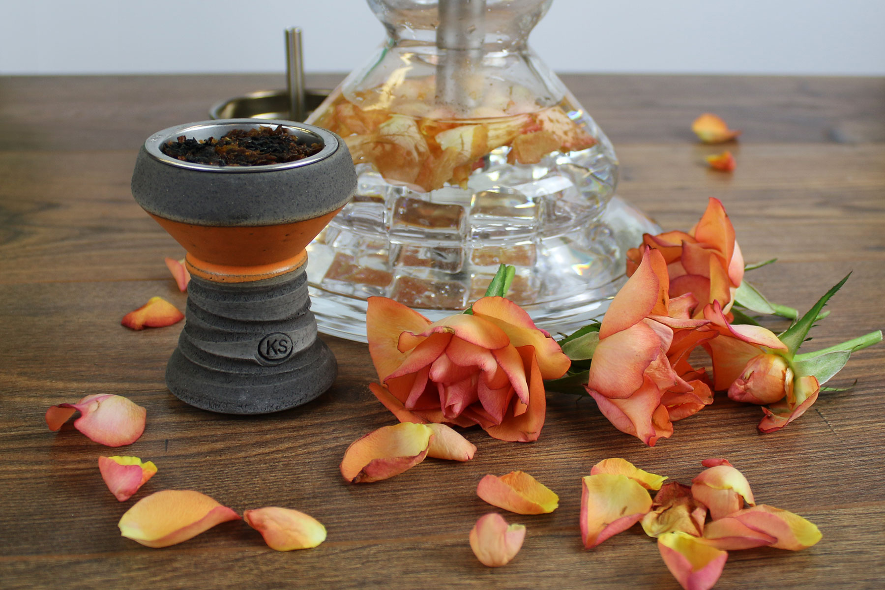 Zusammenstellung eines aufgerauchten Tabakkopfes und einem Shishaglas gefüllt mit Rosenblättern auf einem Holztisch umgeben von Rosenblüten. Die Blüten in der Bowl sehen welk und mitgenommen aus.